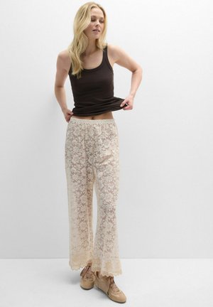 Mujer rubia con top negro sin mangas, pantalones anchos beige con encaje floral y zapatillas beige, posando sobre un fondo blanco liso.