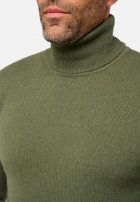 Homme barbu portant un col roulé en tricot vert, montrant le cou et le haut de la poitrine.