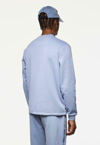 Persona con trenzas, vistiendo un sudadera azul claro, pantalones a juego y una gorra azul claro, de pie de espaldas sobre un fondo liso.