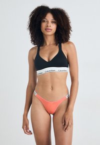 Zwarte bralette met Calvin Klein-logo band, gecombineerd met een koraalkleurige thong met logo-accenten. Zachte stof, minimalistisch ontwerp en contrasterende kleuren.