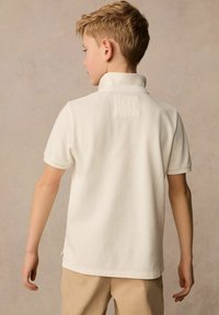 Vit pikétröja med krage, korta ärmar och en fyrkantig tygbit på baksidan. Texturen verkar mjuk och stickad. Bärs med beiga byxor.