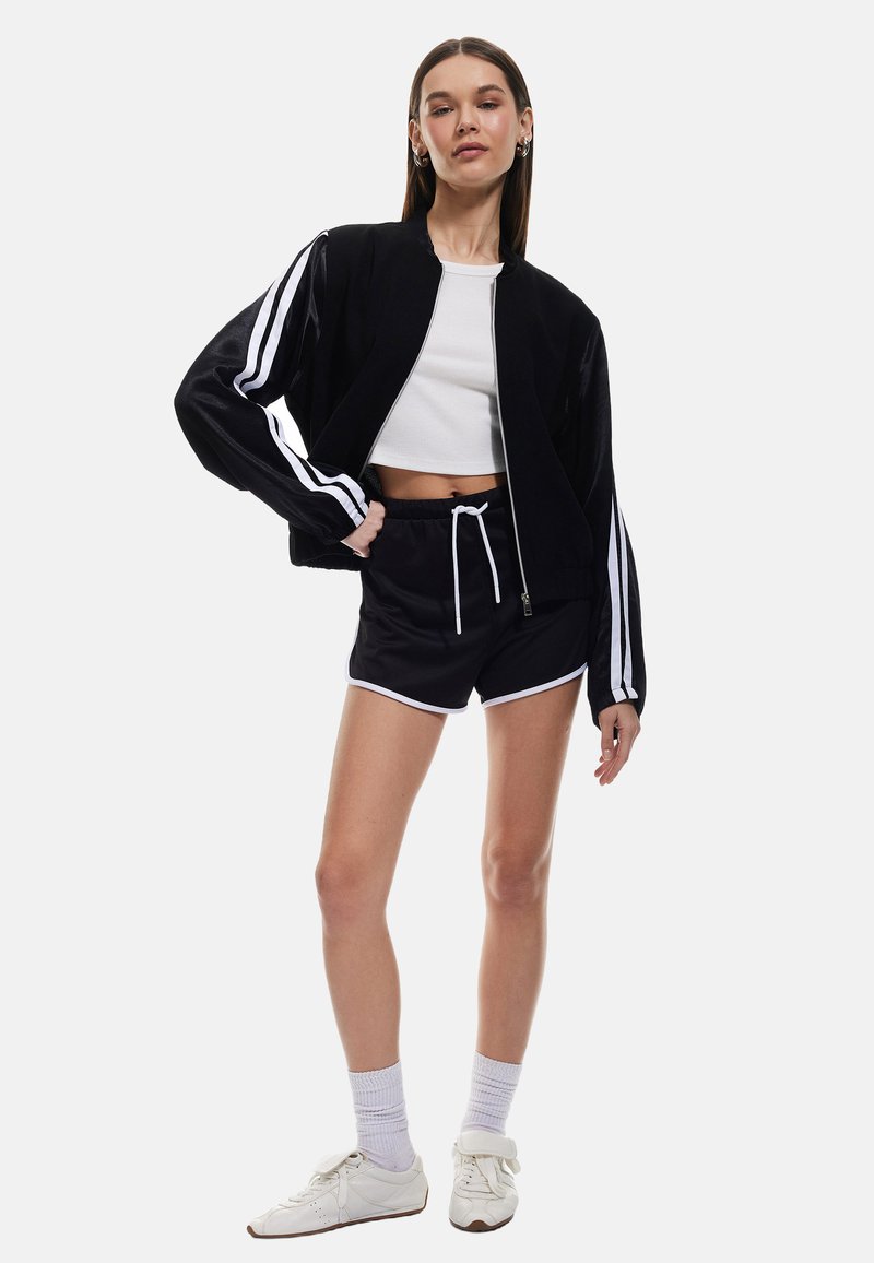 Czarna kurtka bombowca z białymi paskami, w zestawieniu z czarnymi szortami z białymi wykończeniami. Model ma na sobie białe sneakersy i prążkowany top z krótkim rękawem.