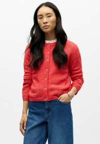 Koraļļu krāsas adīts kardigans ar apaļu izgriezumu, garām piedurknēm un pogām priekšpusē. Valkāts virs baltas topiņas un saskaņots ar zilas džinsa biksēm.