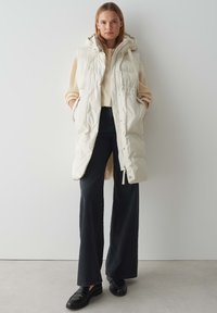 Weißer Puffer-Weste mit Kapuze, ausgestattet mit einem Reißverschluss, Seitentaschen und einem gesteppten Design, getragen über einem beigen Pullover und schwarzen Hosen.