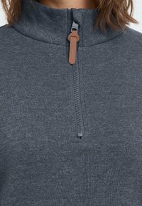 Grijze pullover met een hoge kraag, voorzien van een korte rits en een bruine trekband. De stof heeft een gladde, gestructureerde uitstraling.