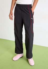 Zwarte sportbroeken met rode bies aan de zijkant, elastische tailleband, zijzakken en een klein grijs logo; gedragen met roze schoenen op een groene ondergrond.