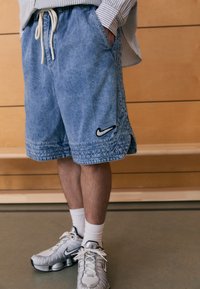 Denim-shortsit haalistuneen sinisellä värillä, joustavalla vyötärönauhalla ja kiristysnauhalla; vasemmassa alareunassa musta Nike-logo. Yhdistetty hopeisiin Nike-tennareihin.