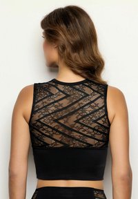 Donna con capelli castani ondulati che indossa un top corto senza maniche di colore nero, con un retro in pizzo trasparente decorato da motivi a zigzag.