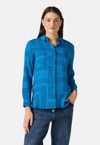 Camisa azul de botones con un patrón geométrico de líneas negras, mangas largas y un bolsillo frontal. Combinada con jeans de mezclilla.