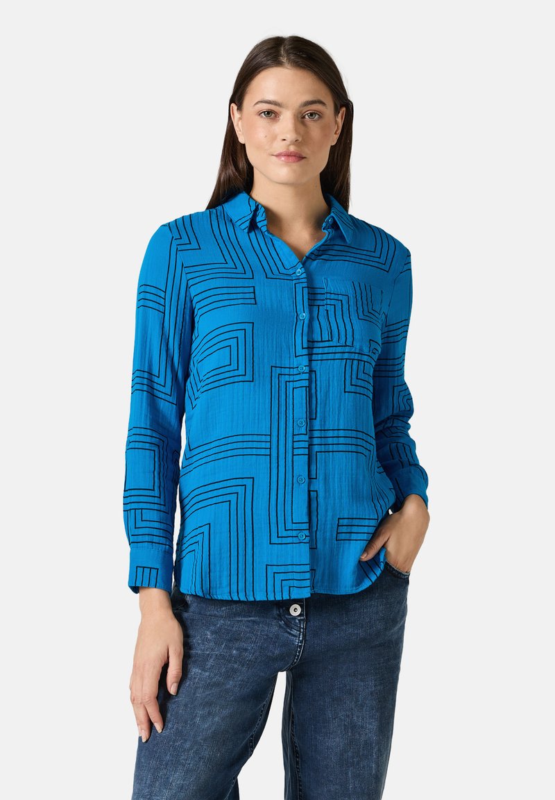 Camisa azul de botones con un patrón geométrico de líneas negras, mangas largas y un bolsillo frontal. Combinada con jeans de mezclilla.