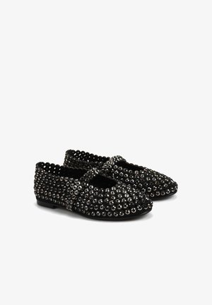 Czarne płaskie buty ozdobione okrągłymi srebrnymi ćwiekami, z falistymi krawędziami i paskiem zapinanym na krzyż. Teksturowana powierzchnia, niska podeszwa.