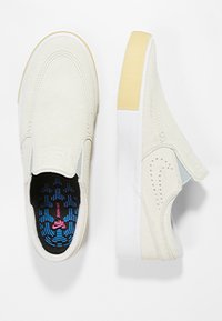 Chaussures sans lacets en daim beige avec une semelle en caoutchouc blanche et un talon accentué jaune. La semelle intérieure présente un motif bleu et noir avec un logo rose.