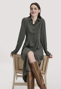 Frau in einem dunkelgrünen, knopfartigen Kleid und braunen, kniehohen Stiefeln, die seitlich auf einem geflochtenen Holzstuhl sitzt, lächelt und nach links schaut.