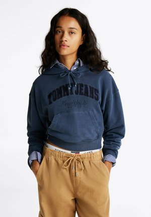 Felpa blu navy con cappuccio e cordini, con la scritta "TOMMY JEANS New York" in rilievo, abbinata a pantaloni beige e una camicia a righe con colletto.
