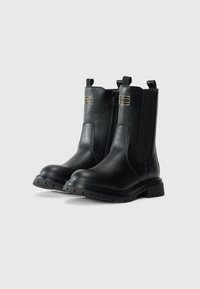 Bottes noires en cuir à la cheville avec fermeture éclair latérale, panneau élastique, languettes et logo rectangulaire doré à l'avant extérieur.