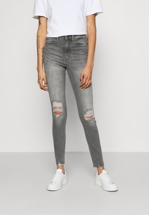 Person trägt hoch taillierte graue Skinny-Jeans mit Knie-Rissen, weiße Turnschuhe und ein einfaches weißes Kurzarmshirt vor neutralem Hintergrund.