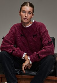 Pflaumenfarbener Pullover mit strukturiertem Strickmuster und schwarzen Bandakzenten an den Ärmeln. Weißer Rüschenkragen lugt hervor. Schwarze Hose.