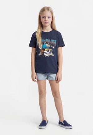 Dziewczyna o długich blond włosach nosi granatowy T-shirt z grafiką czaszki i niebieską czapkę, jasne dżinsowe szorty oraz granatowe wsuwane buty, stoi prosto.