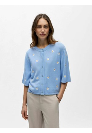 Femeie purtând un cardigan bleu deschis cu nasturi, decorat cu modele mici de flori albe și galbene, și pantaloni bej, stând cu o mână în buzunar.