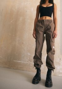 Cargobroek in bruine stof met elastische tailleband, twee zijzakken en een geborduurd logo-embleem. Draagt met zwarte veterschoenen.