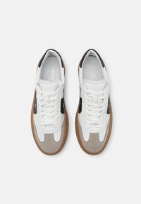 Valkoiset ja harmaat sneakerit, joissa on sileä nahkapinta, mokkanahka-yksityiskohtia ja kumipohja. Varustettu mustilla yksityiskohdilla ja tasaisilla valkoisilla nauhoilla.