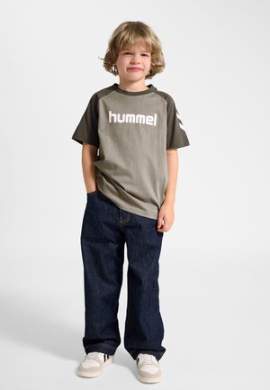 Ung dreng med blondt, krøllet hår iført en grå og sort "hummel" t-shirt, mørkeblå jeans og hvide sneakers, stående med den ene hånd i lommen.