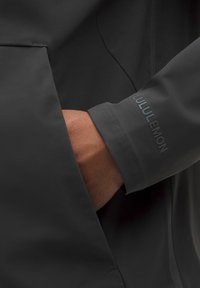 Hand i svart jackficka, synlig ärm med "Lululemon"-logotyp präglad nära handleden.