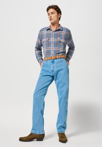 Man wearing a blue plaid shirt tucked into light blue jeans with a tan belt and brown suede boots, standing with hands in pockets.