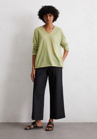 Maglione verde chiaro a maniche lunghe con scollatura a V abbinato a pantaloni ampi neri e sandali neri con dettagli dorati. Tessuto strutturato visibile.