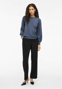 Suéter azul texturizado con dobladillo en forma de concha, combinado con pantalones de pierna ancha negros y tacones de punta negra. Cabello rizado, fondo neutro.