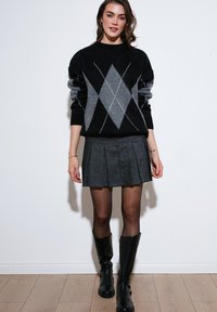Zwart en grijs gebreid vest met diamanten patroon, gecombineerd met een geplooide grijze rok, zwarte maillots en kniehoge zwarte laarzen.