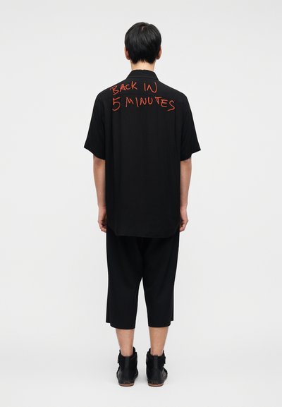 Μαύρο πουκάμισο σε υπερμεγέθη γραμμή με κοντά μανίκια, με κόκκινο κείμενο «BACK IN 5 MINUTES» στην πλάτη. Φορεμένο με μαύρο παντελόνι cropped και μπότες.