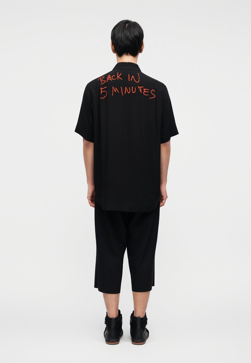 Chemise noire surdimensionnée à manches courtes, avec un texte rouge "BACK IN 5 MINUTES" au dos. Portée avec un pantalon noir raccourci et des bottes.