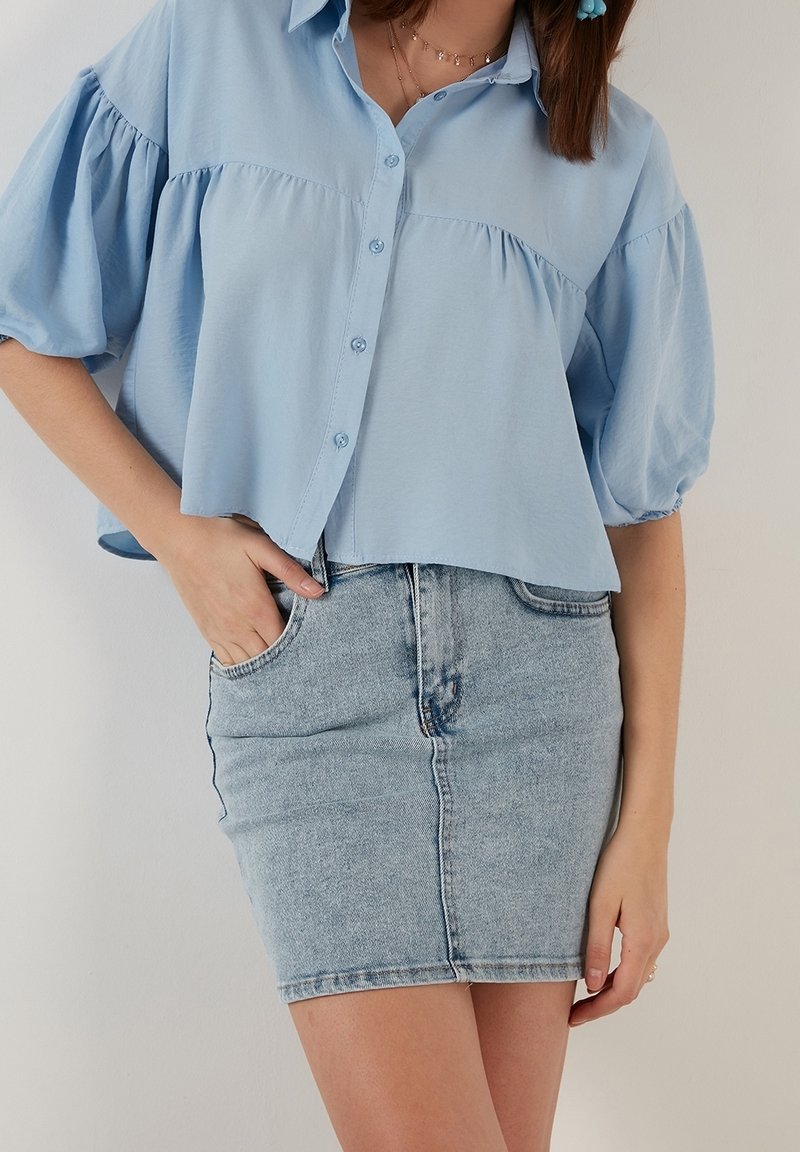 Camisa abotonada azul claro con mangas abullonadas, combinada con una falda de mezclilla ajustada. La tela parece suave, creando una silueta relajada.
