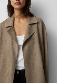 Mujer vestida con un abrigo de ante en color topo sobre una camiseta sin mangas blanca de canalé y jeans negros de tiro alto, de pie contra un fondo claro y liso.
