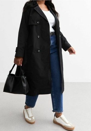 Mujer con gabardina negra, top blanco, vaqueros azules, zapatillas beige y blancas, llevando un bolso negro, caminando sobre un suelo gris.