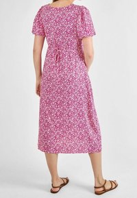 Blumenkleid in Midi-Länge mit kurzen Puffärmeln, tailliertem Bund, rosafarbenem Hintergrund mit weißen Mustern und geradem Saum. Braune Sandalen ergänzen das Outfit.