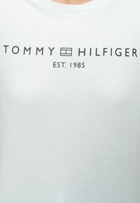 Λευκό μπλουζάκι με μαύρο κείμενο "Tommy Hilfiger" και ένα μικρό τετράγωνο λογότυπο ανάμεσα στις λέξεις, "EST. 1985" τυπωμένο από κάτω.