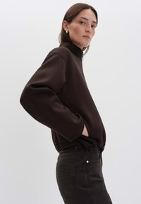 Sweat-shirt en polaire marron avec col montant et zip. Il présente des épaules tombantes et des poches latérales, associé à un jean en denim noir.
