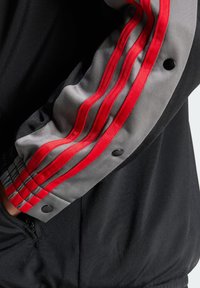 Chaqueta negra con mangas grises y tres rayas rojas, acentuada con botones a presión negros. La tela parece suave con un ligero brillo.