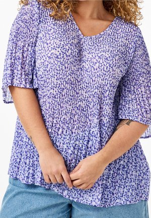 Mujer con cabello rizado que lleva una blusa plisada con estampado floral púrpura y vaqueros azul claro, con las manos juntas a la altura de la cintura.
