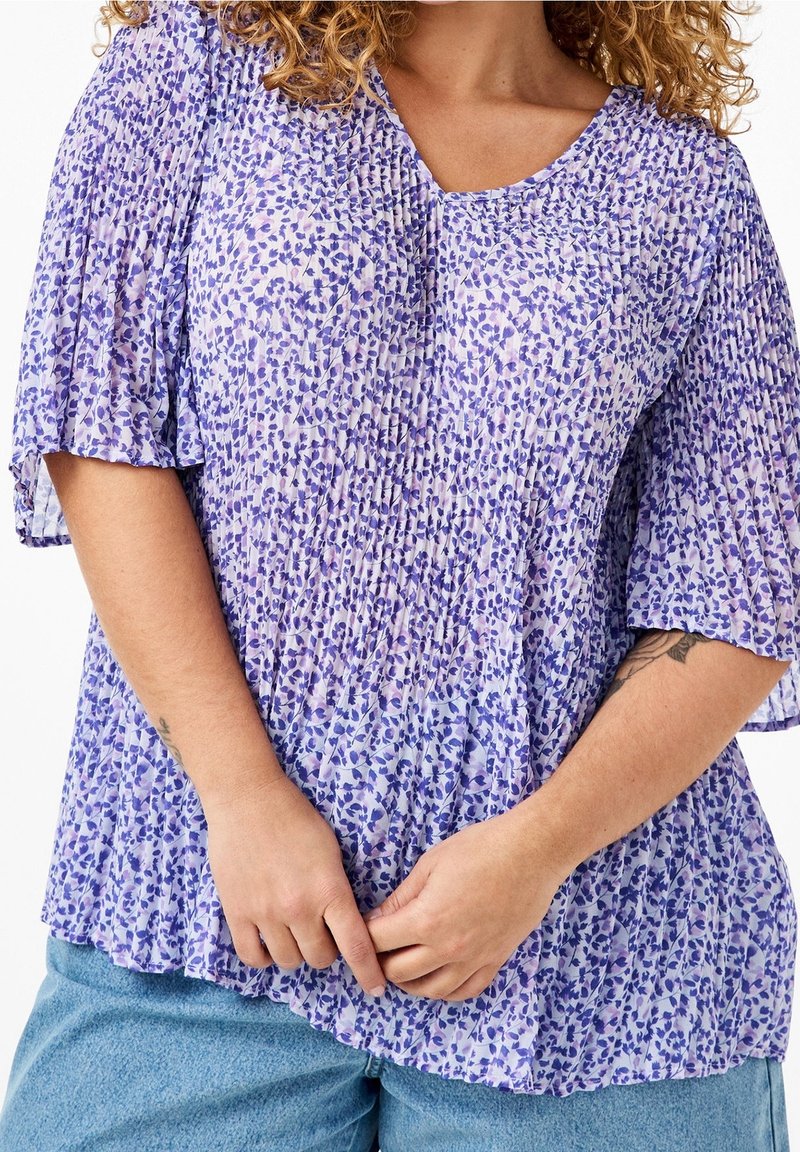 Femme aux cheveux bouclés portant un chemisier plissé à fleurs violet et un jean bleu clair, les mains jointes à la taille.