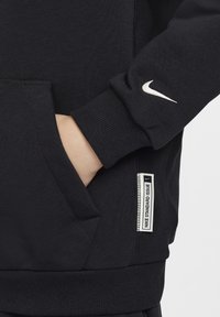 Felpa nera Nike con polsini a costine, tasca a marsupio e logo bianco. Presenta un'etichetta con "NIKE STANDARD ISSUE" in bianco e nero.