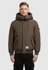 Veste matelassée marron avec une grande capuche, fermeture éclair à l'avant, poches latérales et poignets côtelés. Présente un petit patch logo sur l'ourlet avant.