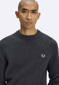 Maglione grigio scuro a coste con scollo tondo, caratterizzato da un piccolo logo ricamato sul lato sinistro del petto. Tessuto morbido e vestibilità comoda.