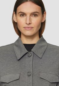 Grijze button-up jas met een kraag, twee borstzakken en een zachte textuur. Voorzien van zwarte knopen en een klassiek ontwerp.