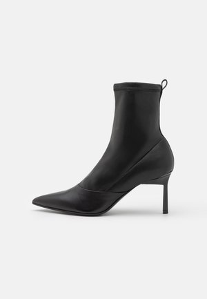 Bottine noire à bout pointu, en cuir lisse et tissu élastique. Elle présente un talon aiguilles fin et une languette à la cheville.