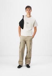 Beige cargo bukser med flere lommer, parret med en let cremefarvet t-shirt med et sort grafisk logo. Accessories inkluderer en sort sling bag.