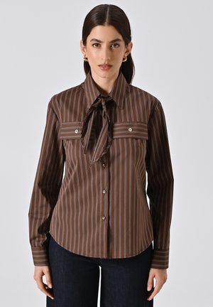 Blusa a righe marroni con colletto a punta, maniche lunghe, dettagli con fiocco al collo e due tasche frontali con accenti di bottone.