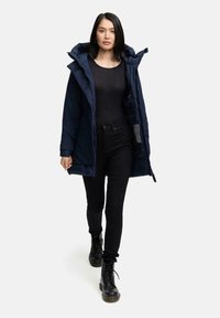 Marineblauer gepolsterter Mantel mit Kapuze, mit einem Reißverschluss vorne und Taschen. Kombiniert mit schwarzen, figurbetonten Hosen und schwarzen Schnürstiefeln.
